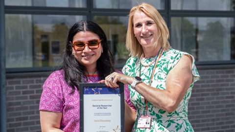 Two people standing in front of a building with large windows; one holds a framed 
